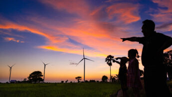 Three people, silhouetted against a sunset, point to a row of wind turbines in Jeneponto Regency, South Sulawesi Province, Indonesia.
