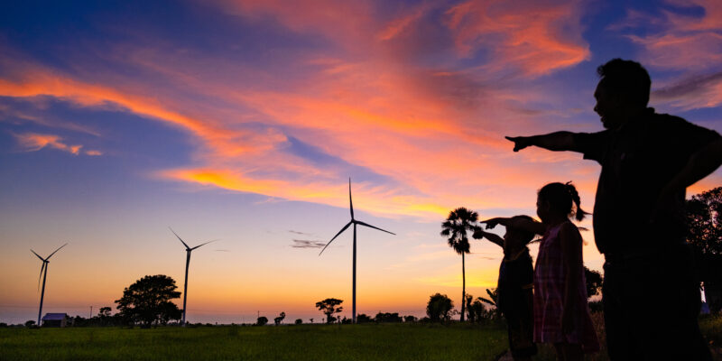 Three people, silhouetted against a sunset, point to a row of wind turbines in Jeneponto Regency, South Sulawesi Province, Indonesia.