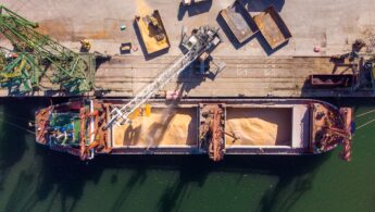 Aerial photo of a cargo ship being unloaded by a crane. Trucks are waiting beside the ship to be loaded.