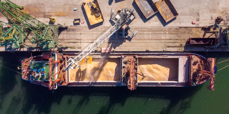 Aerial photo of a cargo ship being unloaded by a crane. Trucks are waiting beside the ship to be loaded.