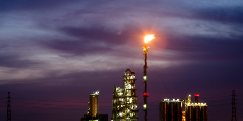 Photograph of gas flaring at twilight. A fire burns at the top of a chimney above a gas plant.