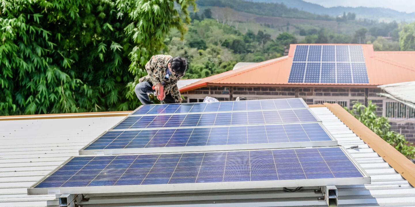Photo of a man installing an array of four solar panels on the roof of a small building. Another roof with solar panels on is visible in the background.
