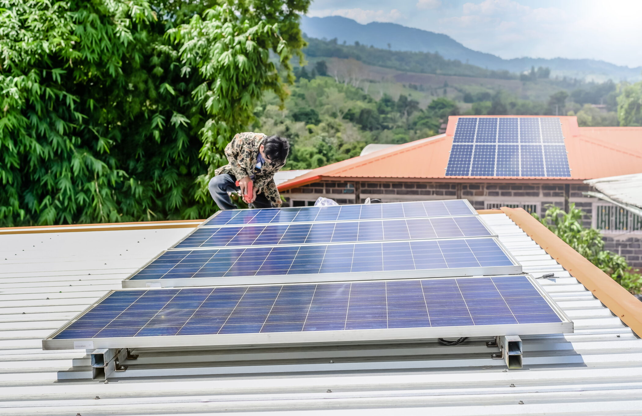 Photo of a man installing an array of four solar panels on the roof of a small building. Another roof with solar panels on is visible in the background.