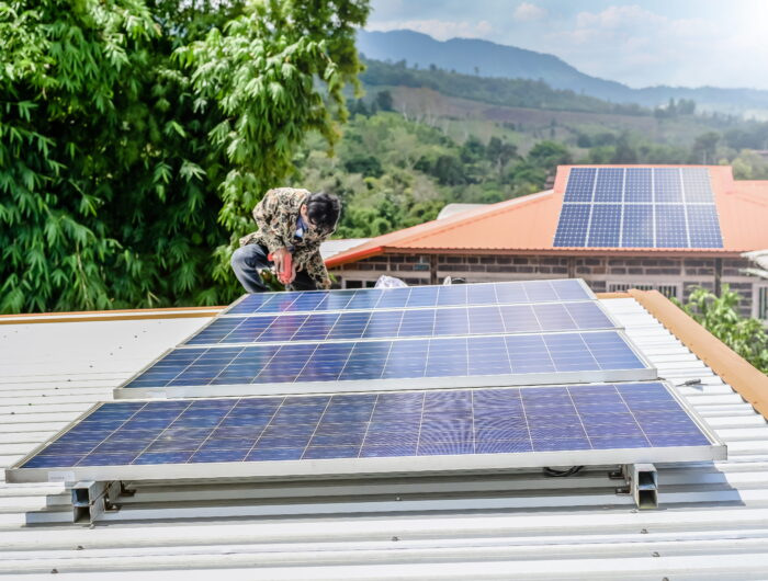 Photo of a man installing an array of four solar panels on the roof of a small building. Another roof with solar panels on is visible in the background.